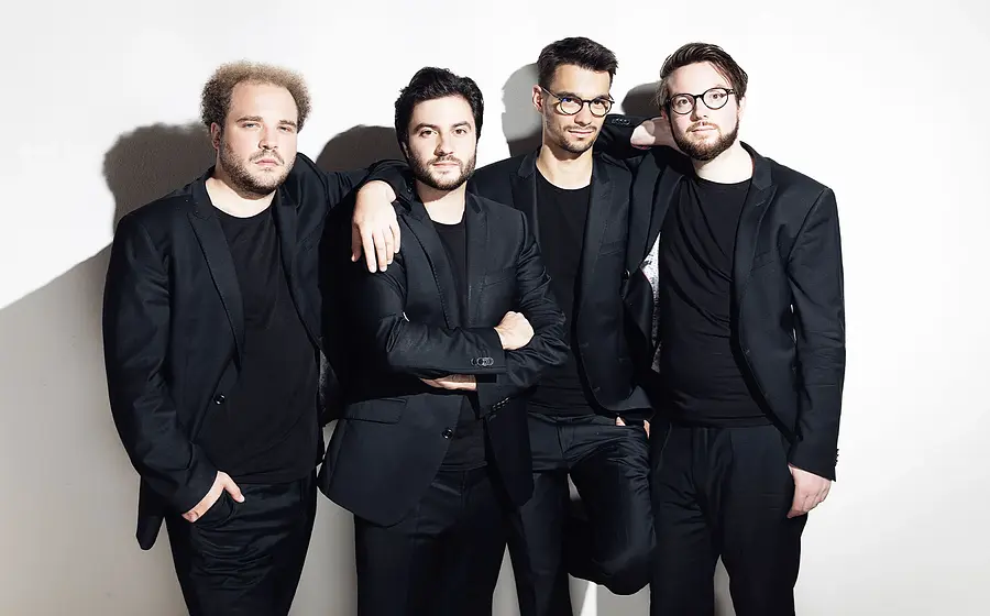 Goldmund Quartett mit vier Musiker:innen in schwarzen Anzügen vor hellem Hintergrund, nebeneinander stehend, teilweise mit verschränkten Armen oder aufgelegten Händen, ohne Instrumente im Bild sichtbar.