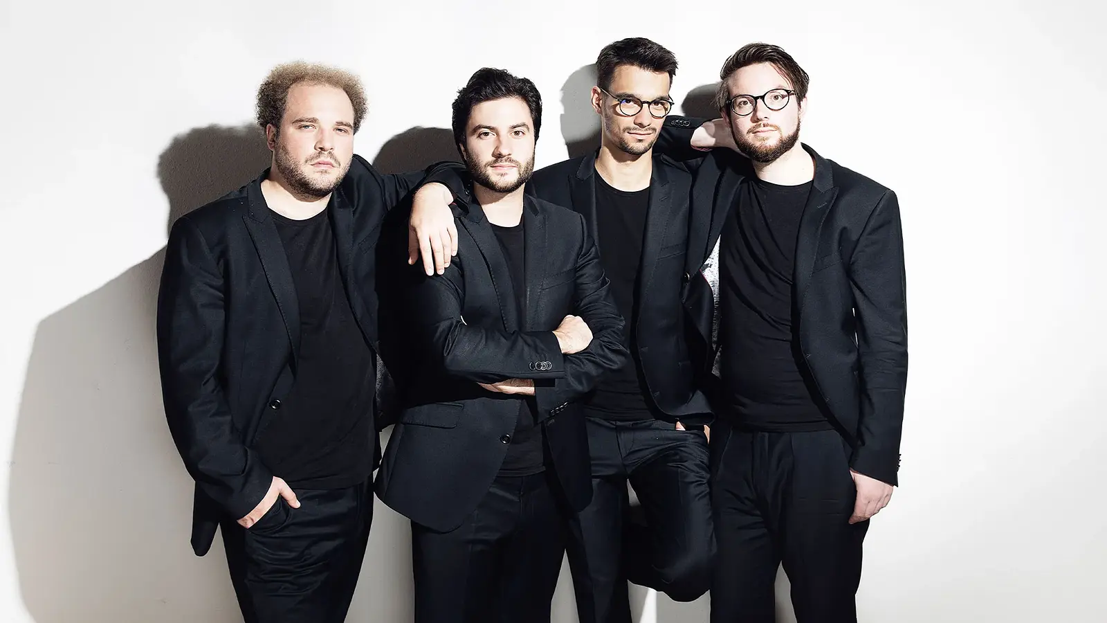 Goldmund Quartett mit vier Musiker:innen in schwarzen Anzügen vor hellem Hintergrund, nebeneinander stehend, teilweise mit verschränkten Armen oder aufgelegten Händen, ohne Instrumente im Bild sichtbar.
