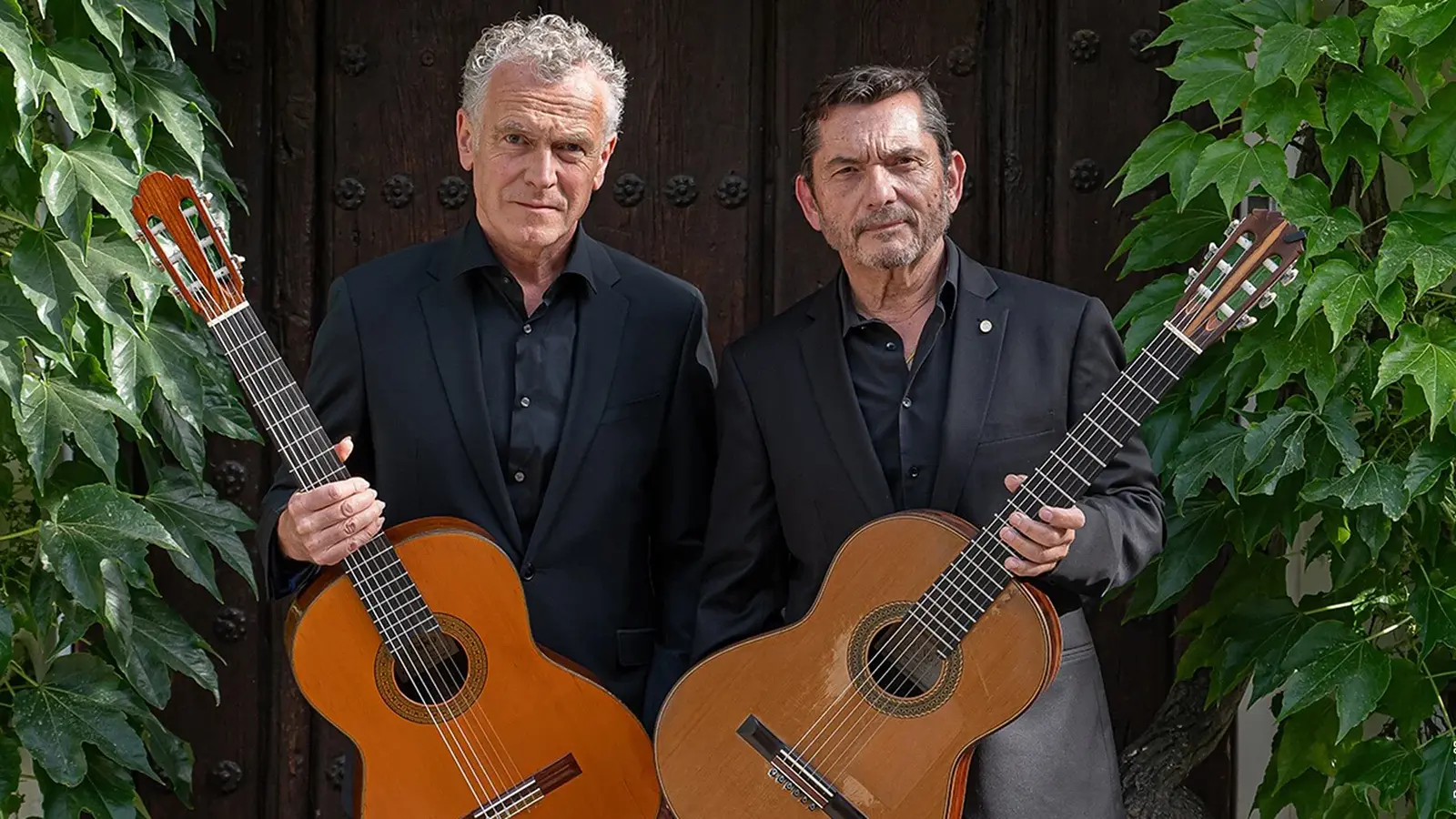 Das Gitarrenduo Wiedermann und GAllardo del Rey mit Gitarren vor einer Holztür, links und rechts im Bild Kletterpflanzen