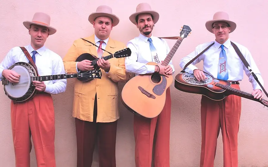 Die vier Mitglieder der Truffle Valley Boys mit Gitarren in farbiger Kleidung und rosa Hüten vor rosa Hintergrund