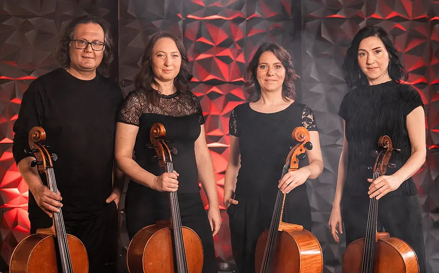 Gruppenporträt eines Celloensembles vor einer geometrisch strukturierten Wand mit roter Beleuchtung. Vier Musikerinnen und Musiker in schwarzer Konzertkleidung stehen nebeneinander und halten ihre Violoncelli vor sich.