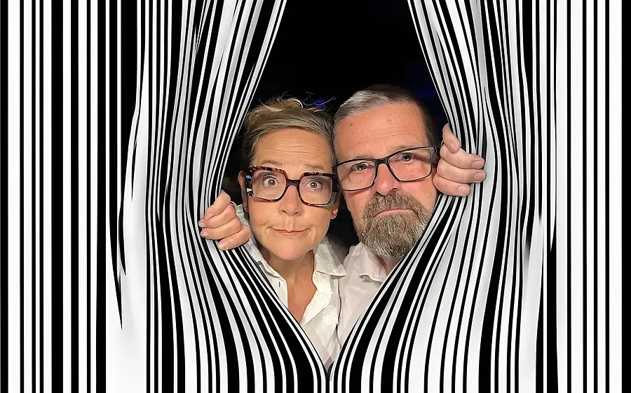 Porträtfoto mit zwei Personen in weißen Hemden und markanten Brillen, die durch eine Öffnung in einem schwarz weißen Streifenmuster eines Strichcodes blicken