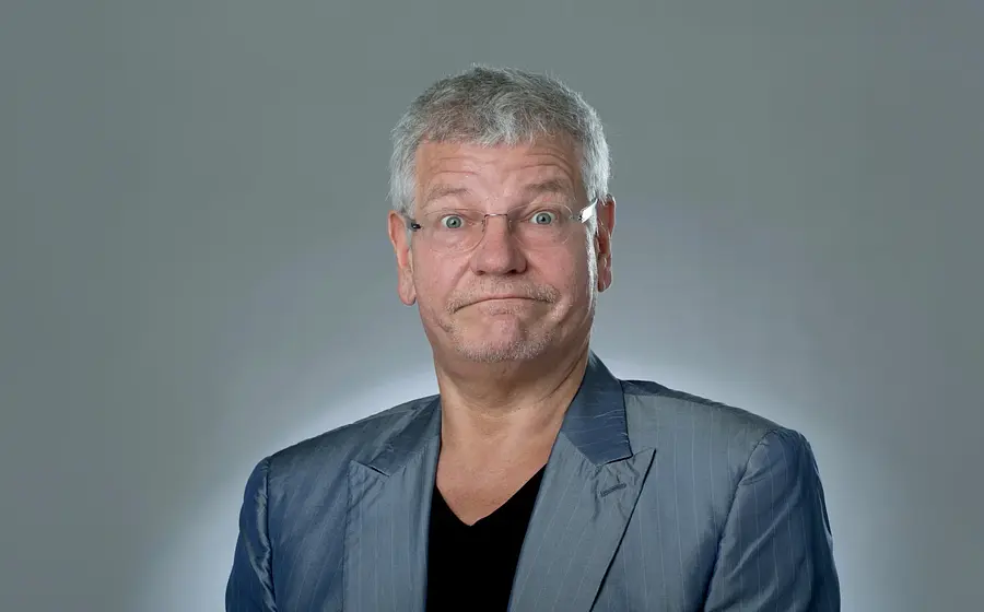 Porträt des Kabarettisten Werner Koczwara mit grauem Haar und Brille in silbernem Sakko vor hellem Hintergrund. Die Person blickt mit weit geöffneten Augen direkt in die Kamera und zieht die Lippen zu einem skeptischen Ausdruck zusammen.