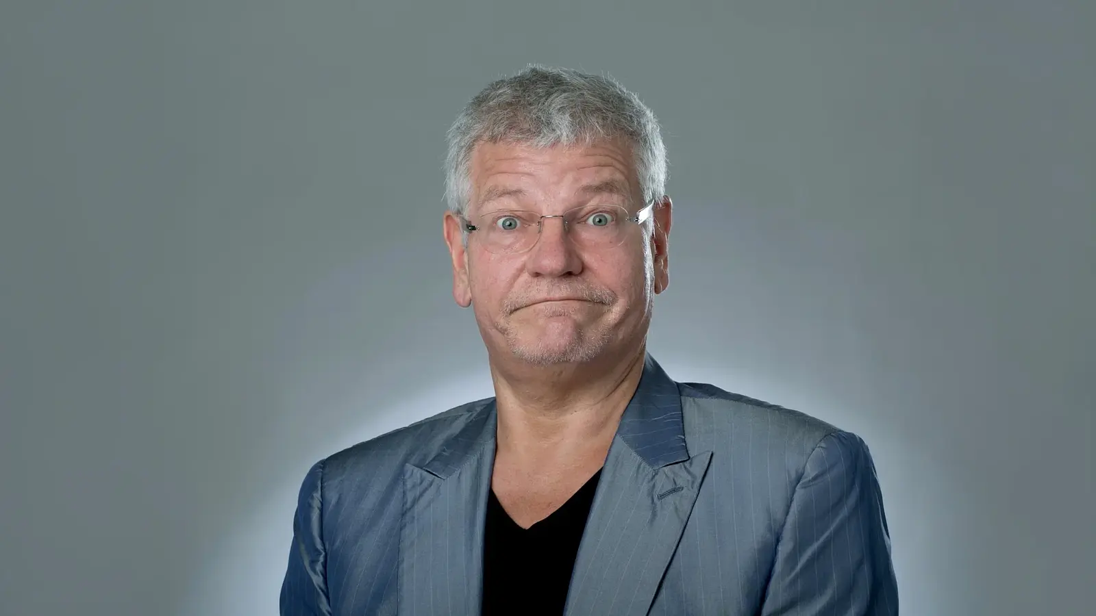 Porträt des Kabarettisten Werner Koczwara mit grauem Haar und Brille in silbernem Sakko vor hellem Hintergrund. Die Person blickt mit weit geöffneten Augen direkt in die Kamera und zieht die Lippen zu einem skeptischen Ausdruck zusammen.