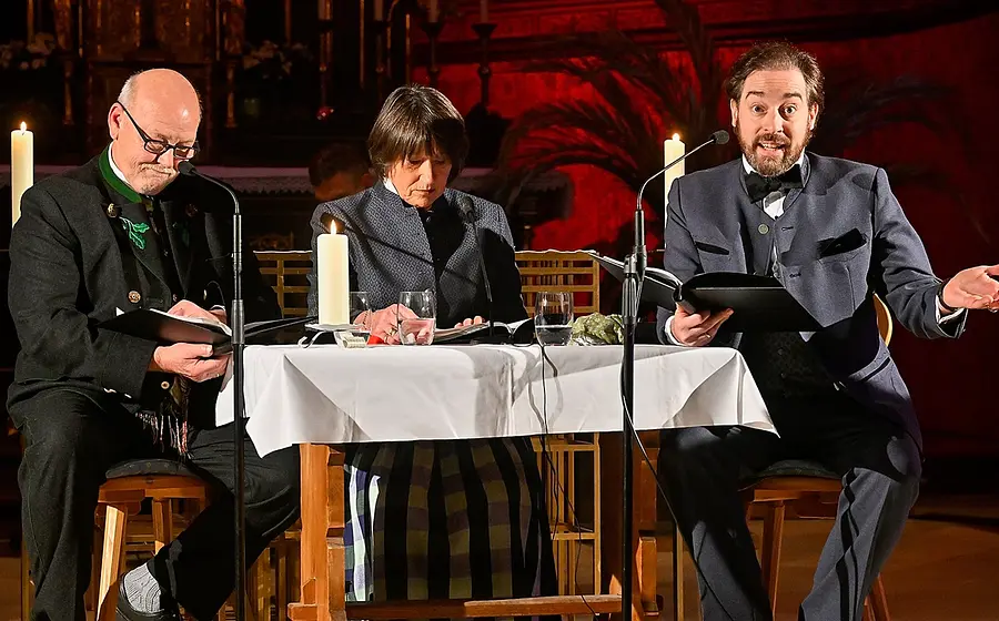 Drei Vortragende sitzen bei einer Lesung an einem Tisch mit Kerzen und Mikrofonen in einem festlich beleuchteten Kirchenraum