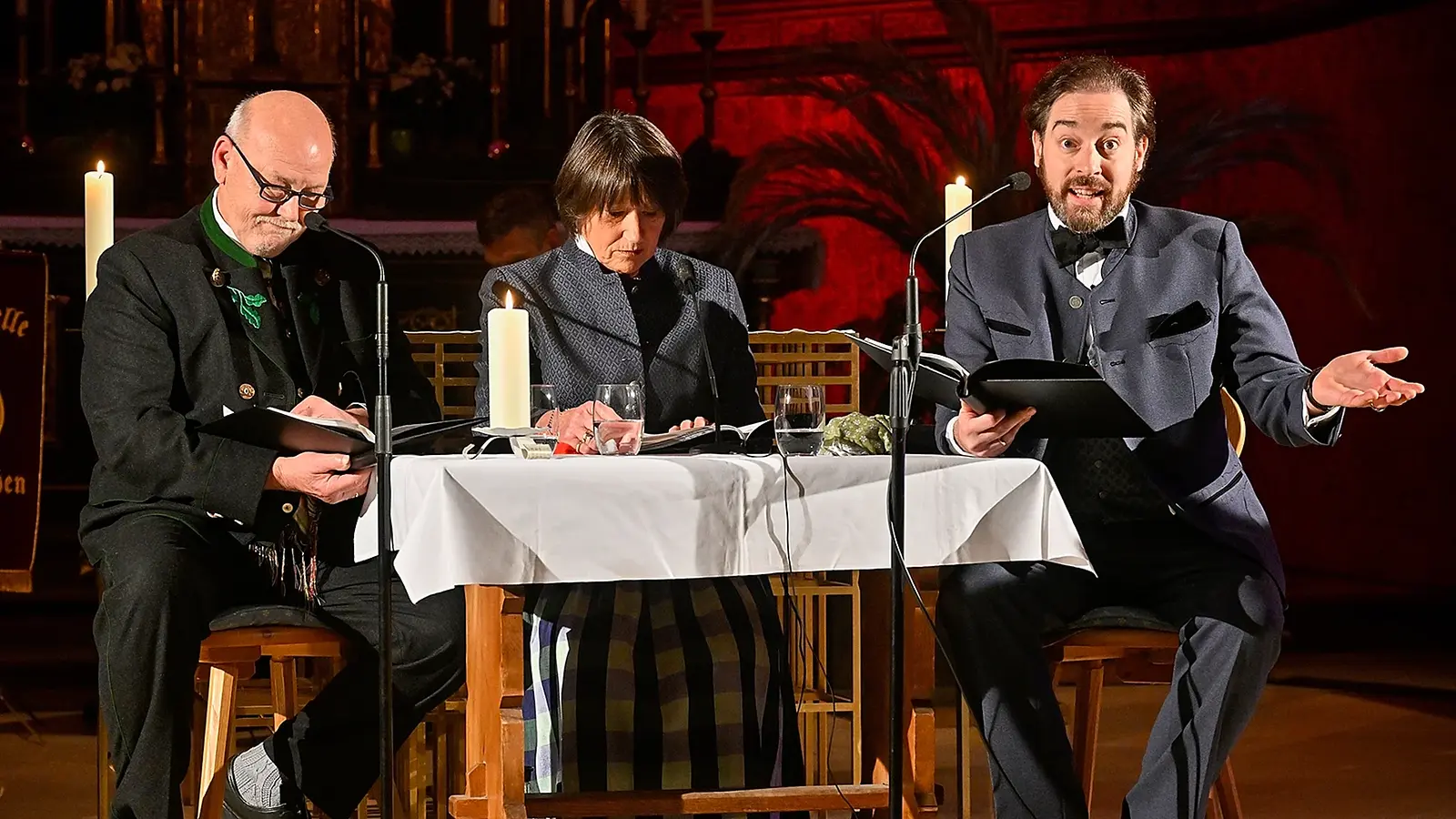Drei Vortragende sitzen bei einer Lesung an einem Tisch mit Kerzen und Mikrofonen in einem festlich beleuchteten Kirchenraum