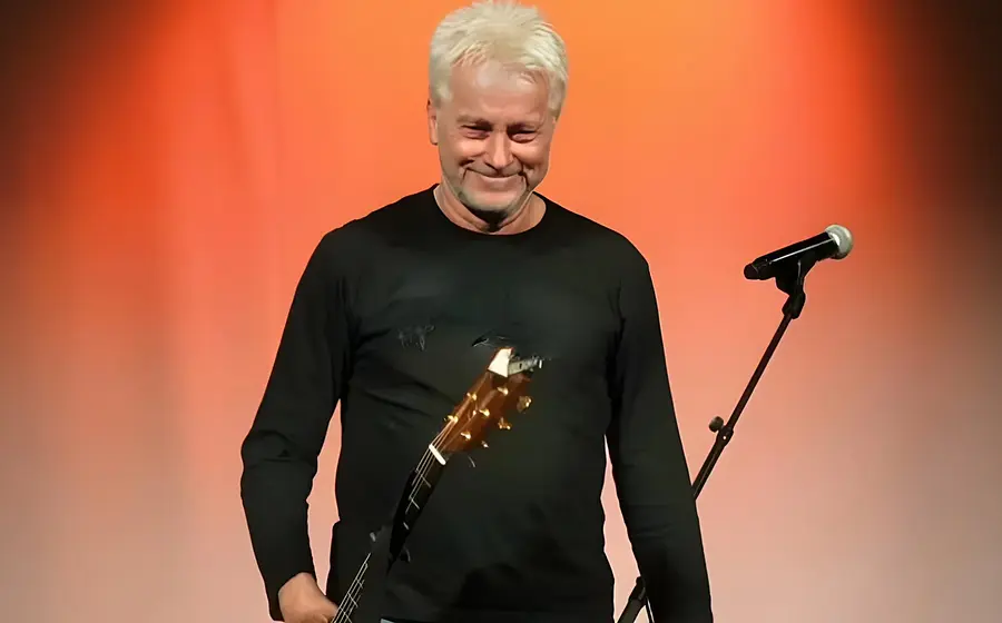 Der Künstler Werner Schmidbauer schwarz gekleidet mit Gitarre und einem Mikrofonständer, der Hintergrund ist orangerot beleuchtet