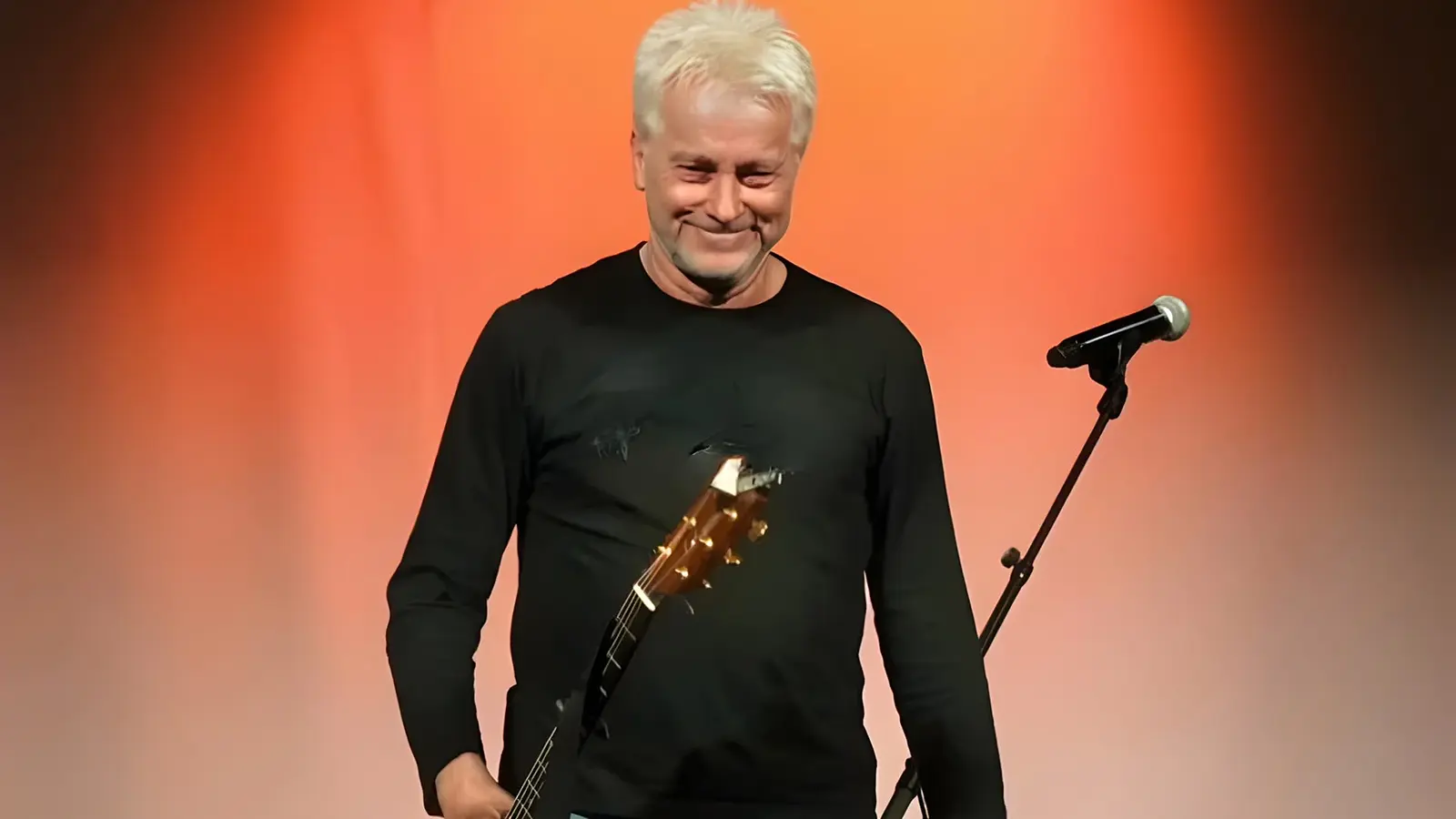 Der Künstler Werner Schmidbauer schwarz gekleidet mit Gitarre und einem Mikrofonständer, der Hintergrund ist orangerot beleuchtet