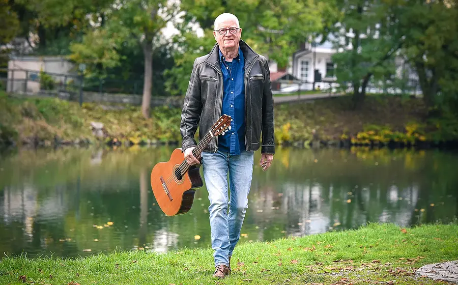 Willy Sommerwerk in Jeans und Lederjacke mit Gitarre auf einer Wiese stehend, im Hintergrund ein Bach und Gebäude