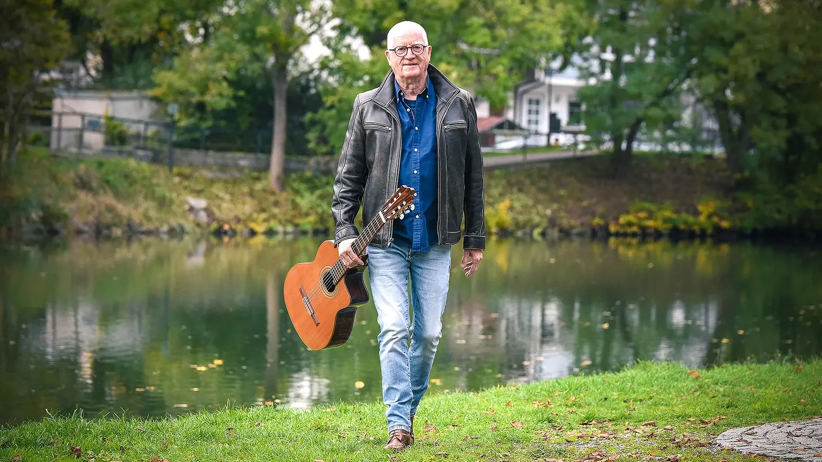 Willy Sommerwerk in Jeans und Lederjacke mit Gitarre auf einer Wiese stehend, im Hintergrund ein Bach und Gebäude