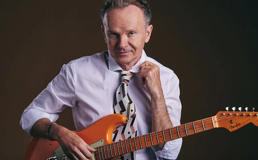 Willy Astor in einem hellblauen Hemd und gemusterter Krawatte mit einer E-Gitarre vor dunklem Hintergrund