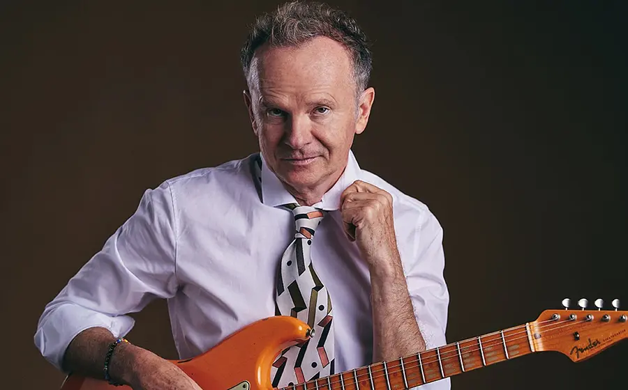 Bild des Künstlers mit weißer Krawatte und weißem Hemd und einer orangefarbenen E-Gitarre