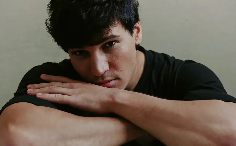 Wincent Weiss in schwarzem T Shirt stützt den Kopf auf verschränkte Arme und blickt direkt in die Kamera, vor hellem Hintergrund