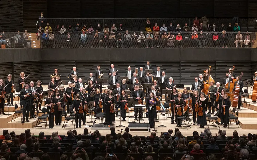Bühnenfoto des Akademischen Sinfonieorchesters München mit großem Orchester auf der Bühne der Isarphilharmonie vor Publikum, Musikerinnen und Musiker stehen nach dem Konzert zwischen Streichern, Bläsern und Kontrabässen, während die Zuschauerränge im Hintergrund besetzt sind.