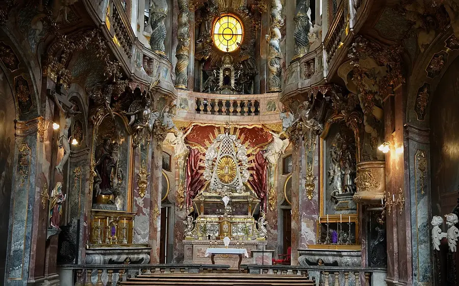 Innenraum der barocken Asamkirche mit reich verziertem Altar, farbigem Marmor, goldenen Ornamenten, Skulpturen und einem großen Rundfenster im oberen Bereich, frontal aufgenommen mit Blick auf den Hauptaltar.