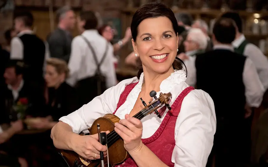 Person in Dirndl hält eine Violine in den Händen und steht in einem Wirtshaus mit weiteren Personen im Hintergrund, die in traditioneller Kleidung und unscharf im Raum verteilt sind.