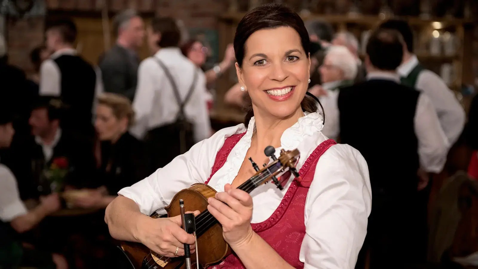 Person in Dirndl hält eine Violine in den Händen und steht in einem Wirtshaus mit weiteren Personen im Hintergrund, die in traditioneller Kleidung und unscharf im Raum verteilt sind.