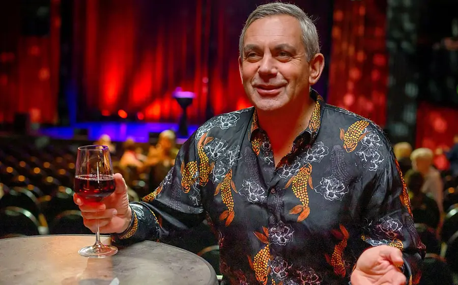 Porträtfoto von Wladimir Kaminer an einem Stehtisch mit einem Glas Rotwein in der Hand, vor einem Veranstaltungsraum mit Reihen von Stühlen und rot beleuchteter Bühne im Hintergrund