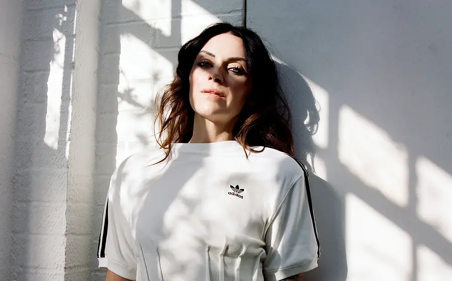 Porträtfoto von Amy Macdonald in weißem T-Shirt vor heller Wand, mit langen dunklen Haaren und Sonnenlicht, das durch Fensterrahmen Schatten auf Gesicht und Kleidung wirft