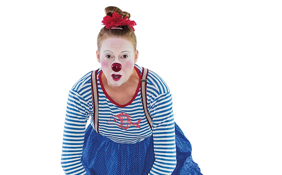 Porträtfoto mit Clownfigur mit weiß geschminktem Gesicht, roter Nase und blau weiß gestreiftem Oberteil, die sich nach vorne beugt, vor hellem neutralem Hintergrund
