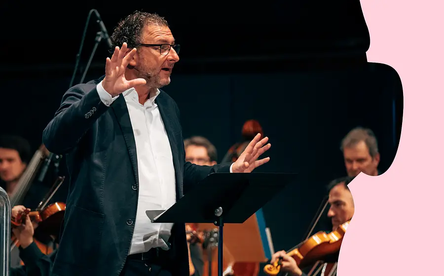 Person am Rednerpult vor einem Orchester auf einer Konzertbühne, umgeben von rosafarbenen grafischen Elementen am Bildrand.
