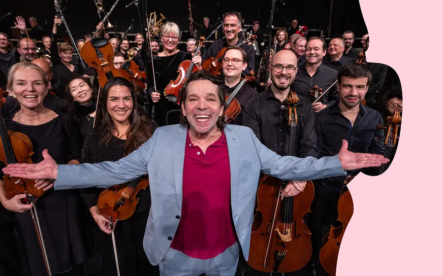 Person mit ausgebreiteten Armen vor einem Orchester; die Musikerinnen und Musiker halten Streich- und Blasinstrumente und blicken in die Kamera.