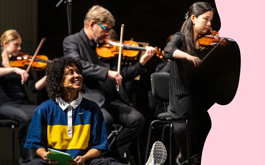 Person mit Mikrofon sitzt vor einem Streichensemble auf der Bühne; im Hintergrund spielen mehrere Musikerinnen und Musiker mit Violinen.