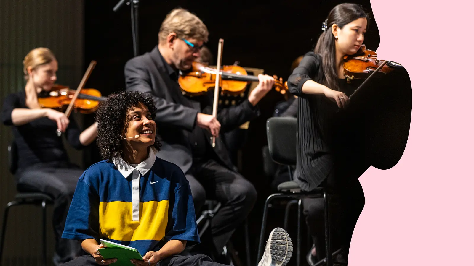 Person mit Mikrofon sitzt vor einem Streichensemble auf der Bühne; im Hintergrund spielen mehrere Musikerinnen und Musiker mit Violinen.