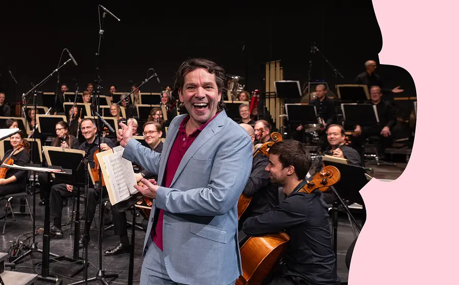 Person mit geöffnetem Notenheft steht vor einem Orchester auf der Bühne und gestikuliert in Richtung der Musikerinnen und Musiker.