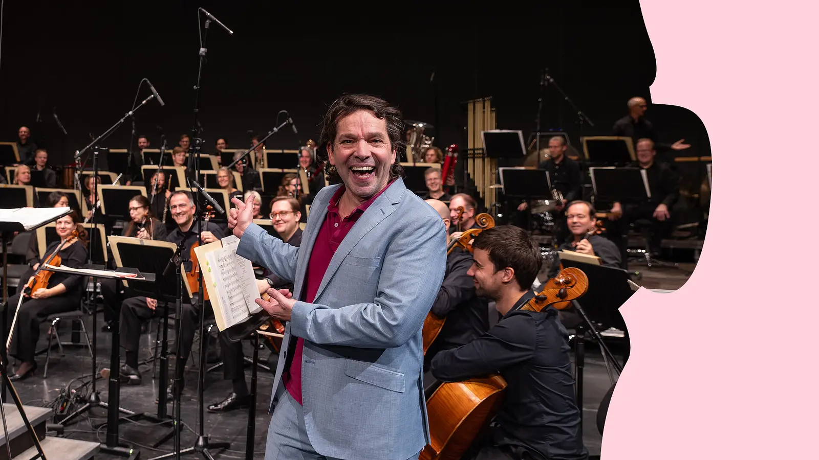 Person mit geöffnetem Notenheft steht vor einem Orchester auf der Bühne und gestikuliert in Richtung der Musikerinnen und Musiker.