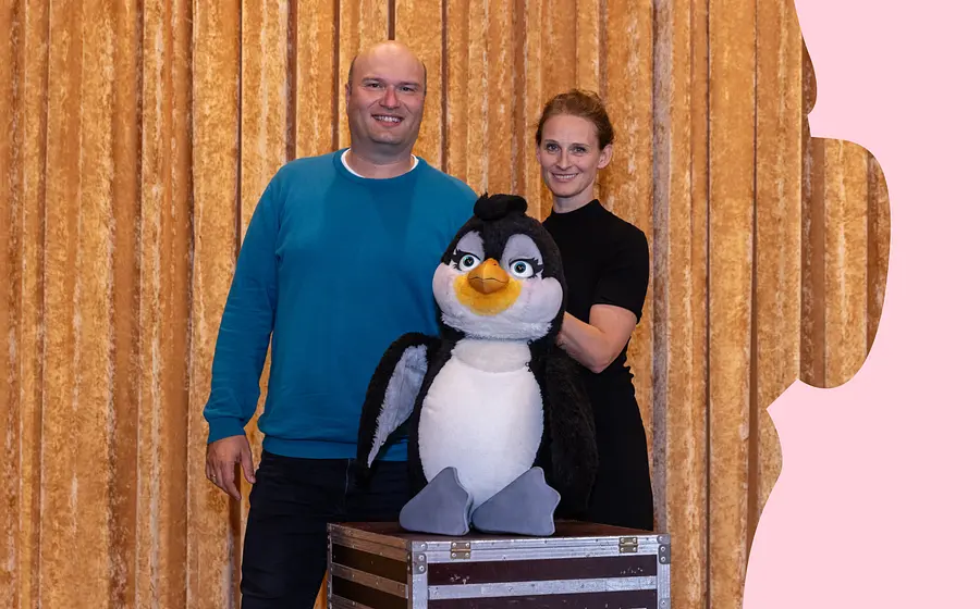 Zwei Personen stehen mit einer großen Pinguinfigur vor einem goldfarbenen Bühnenvorhang. Die Pinguinfigur steht auf einer Transportkiste.
