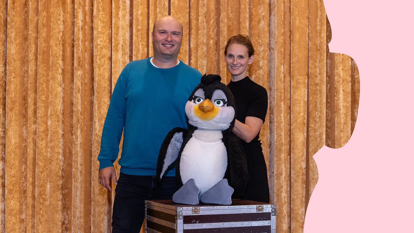Zwei Personen stehen mit einer großen Pinguinfigur vor einem goldfarbenen Bühnenvorhang. Die Pinguinfigur steht auf einer Transportkiste.