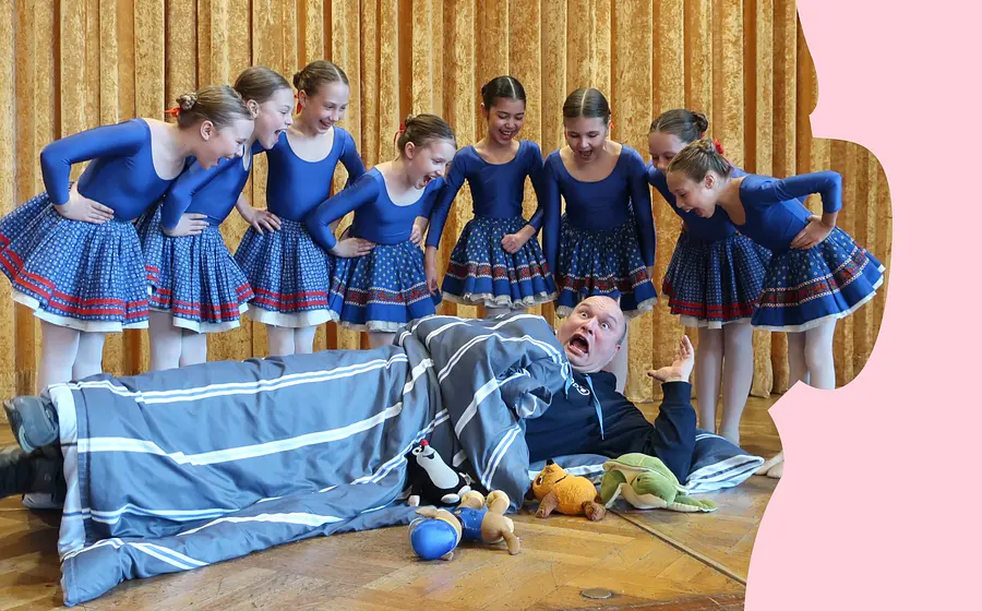 Gruppe von Kindern in blauen Tanzkostümen steht auf einer Bühne um eine Person, die zwischen Bettdecke und Stofftieren auf dem Boden liegt.