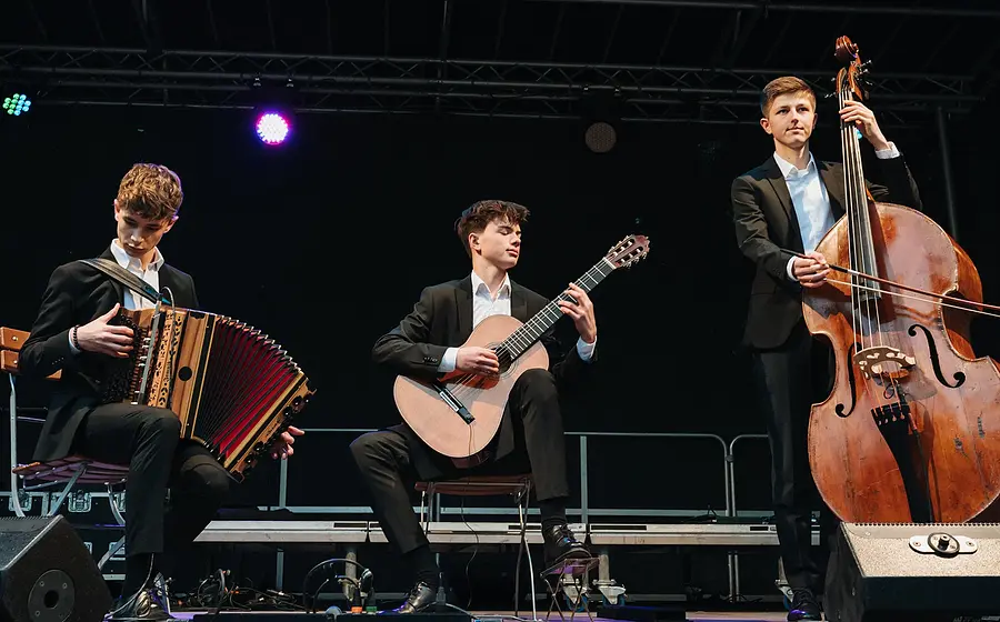 Drei junge Musiker spielen auf einer Bühne Akkordeon, Gitarre und Kontrabass.