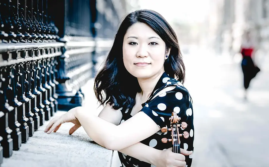 Tianwa Yang in schwarzer Bluse mit weißen Punkten, hält ein Instrument, stützt die Ellbogen auf und blickt in die Kamera