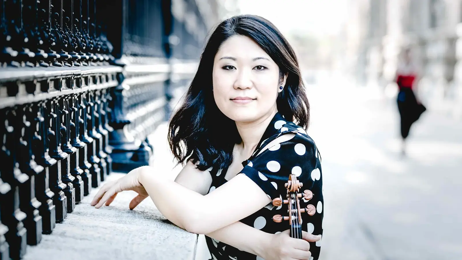 Tianwa Yang in schwarzer Bluse mit weißen Punkten, hält ein Instrument, stützt die Ellbogen auf und blickt in die Kamera