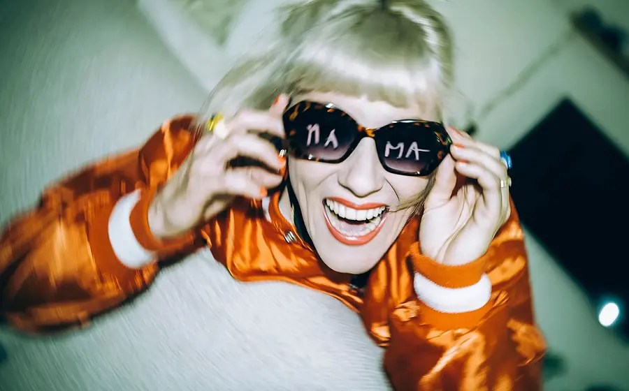 Person mit blondem Haar und Sonnenbrille lächelt in die Kamera und trägt eine glänzende orange Jacke; in den Brillengläsern ist Schrift zu sehen, Aufnahme mit unscharfem Hintergrund und starker Bewegung.