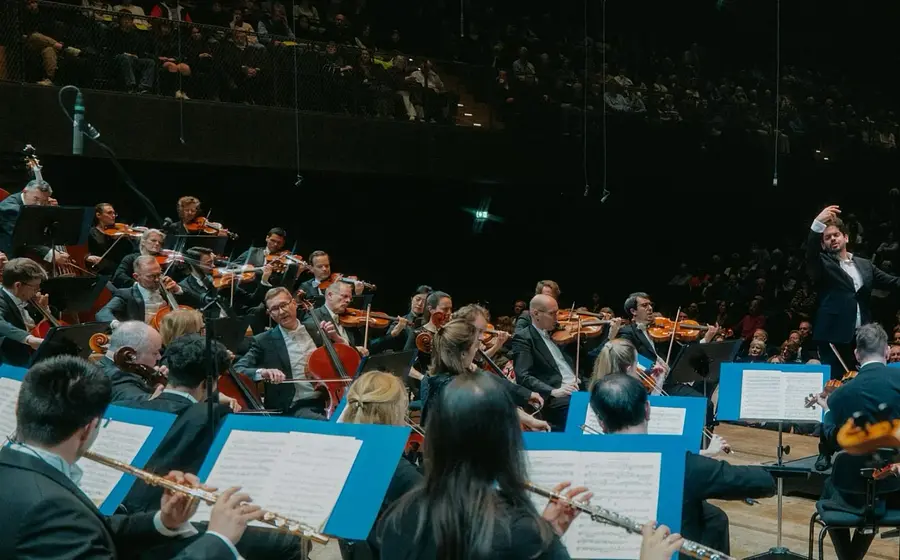Orchester bei einem Konzert mit Dirigent auf der Bühne, vor großem Publikum in einem Konzertsaal.