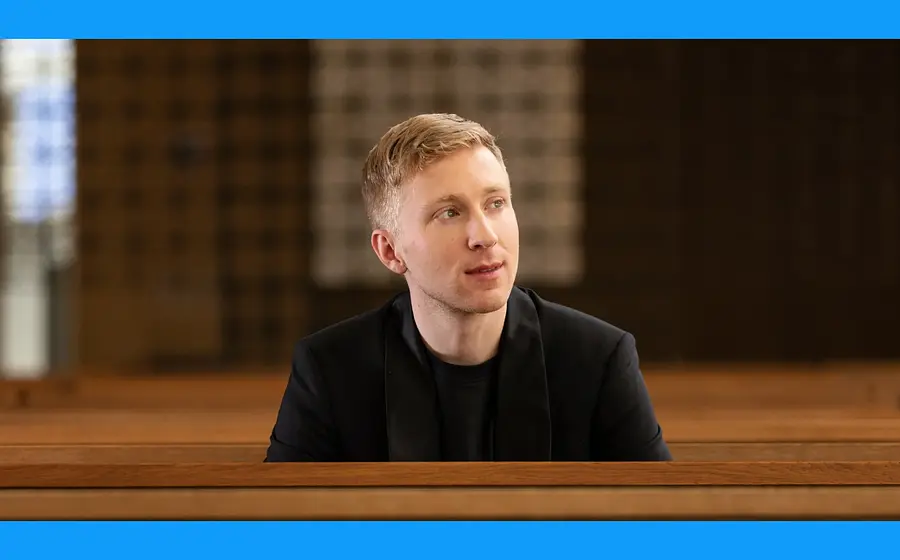 Patrick Hahn an einem Holzpult, zur Seite Blickend, das Bild hat einen blauen Rahmen