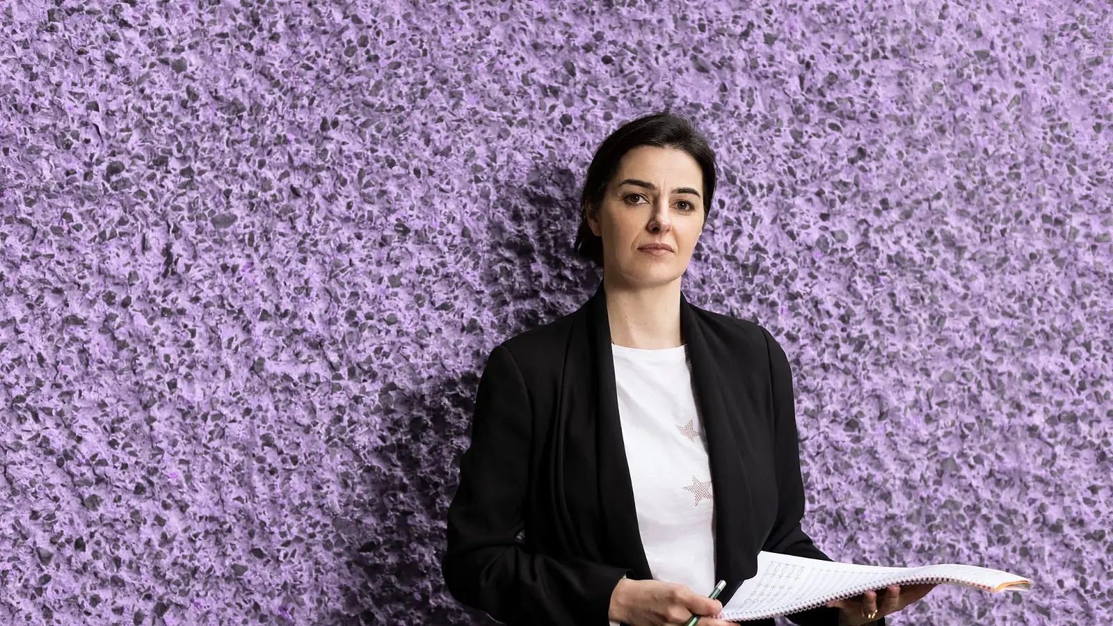 Eine Person in weißem Shirt, schwarzem Blazer mit Notenblatt in den Händen vor violetter rauher Steinwand zur Veranstaltung KOMPONISTENPORTRÄT DOBRINKA TABAKOVA