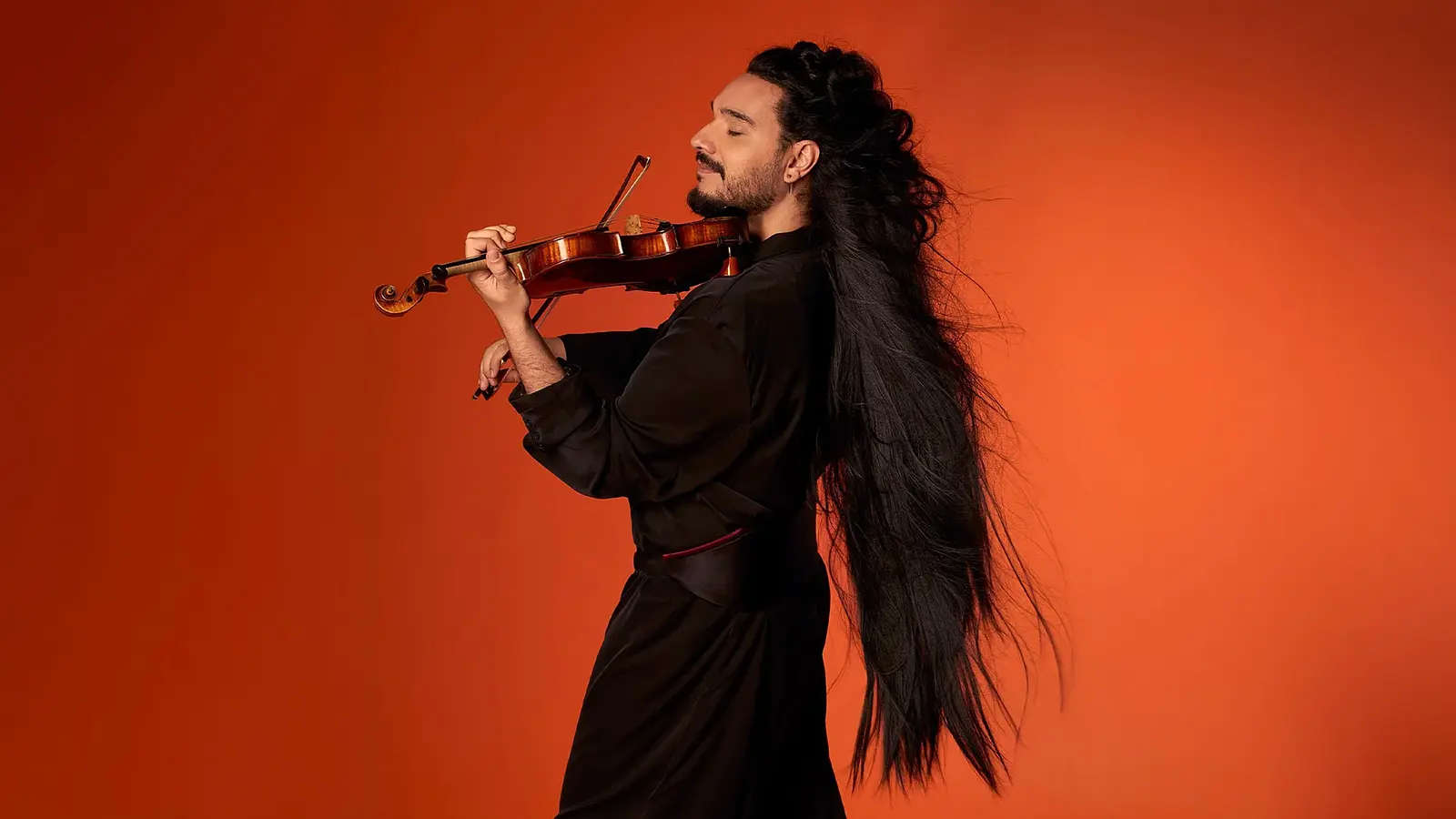 Nemanja Radulovic spielt Violine vor orangefarbenem Hintergrund mit langem, nach hinten fallendem Haar und schwarzer Kleidung.