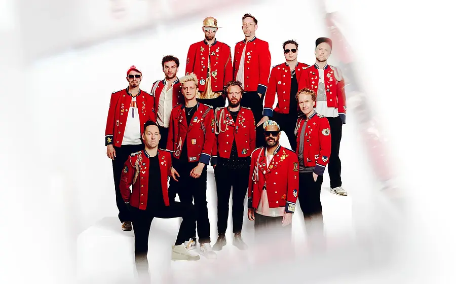 Band MEUTE mit mehreren Musikerinnen und Musikern in roten Uniformjacken steht auf weißen Podesten vor hellem Hintergrund und posiert als Gruppe.