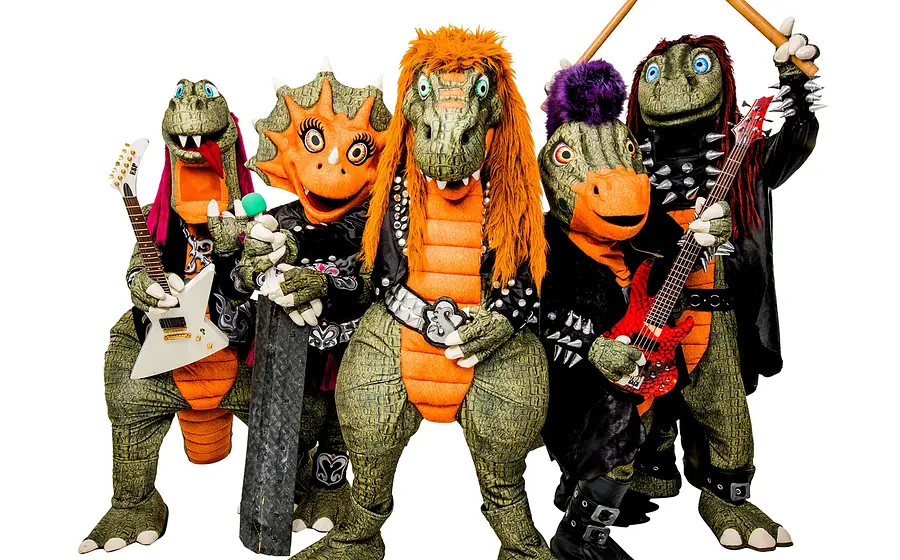 Pressefoto von Heavysaurus mit fünf Dinosaurierfiguren in Rockkostümen vor hellem Hintergrund, darunter E-Gitarren und Drumsticks, mit bunten Haaren, Nieten und schwarzen Bühnenoutfits