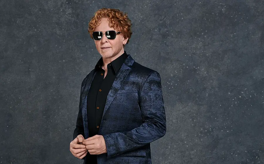 Porträtfoto von Mick Hucknall in dunkelblauem glänzendem Sakko und schwarzem Hemd vor grauem Hintergrund, mit Sonnenbrille und lockigem rotem Haar