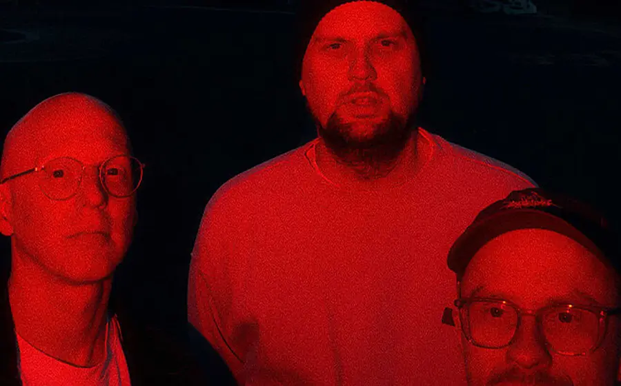 Bandfoto von Smile And Burn mit drei Bandmitgliedern vor schwarzem Hintergrund, in rotes Licht getaucht, zwei Personen mit Brille im Vordergrund und eine Person mit Mütze mittig im Hintergrund.
