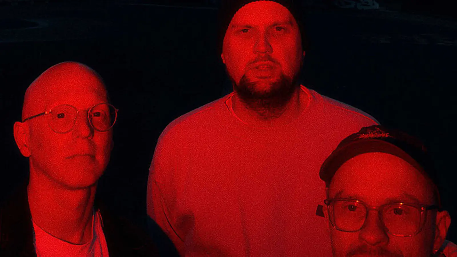 Bandfoto von Smile And Burn mit drei Bandmitgliedern vor schwarzem Hintergrund, in rotes Licht getaucht, zwei Personen mit Brille im Vordergrund und eine Person mit Mütze mittig im Hintergrund.