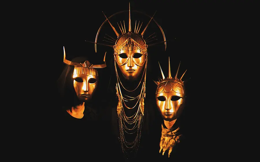 Bandfoto von Imperial Triumphant mit drei Personen in schwarzen Gewändern und goldfarbenen, kunstvoll verzierten Masken vor schwarzem Hintergrund, die mittlere Maske mit strahlenförmigem Kopfschmuck und die beiden seitlichen mit markanten Hörnern beziehungsweise Spitzen