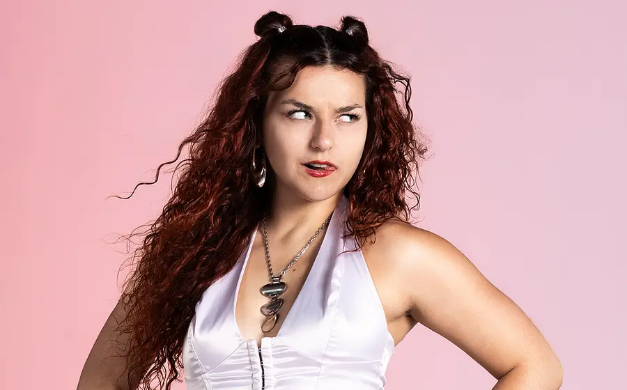 Portrait von Maiva vor hellrotem Hintergrund. Die Künstlerin schaut frech rechts neben der Kamera vorbei und streckt die Zunge raus.