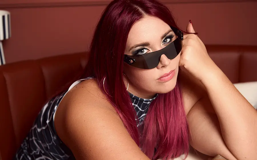 Porträtfoto von PÄM mit langen magentafarbenen Haaren, dunkler futuristischer Sonnenbrille und großen Creolen, auf einer braunen Sitzbank liegend und den Kopf auf die Hand gestützt, vor warm beleuchtetem Hintergrund.