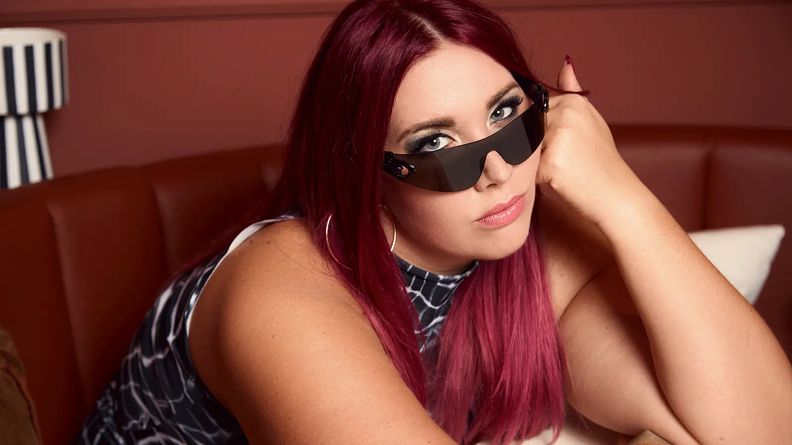 Porträtfoto von PÄM mit langen magentafarbenen Haaren, dunkler futuristischer Sonnenbrille und großen Creolen, auf einer braunen Sitzbank liegend und den Kopf auf die Hand gestützt, vor warm beleuchtetem Hintergrund.
